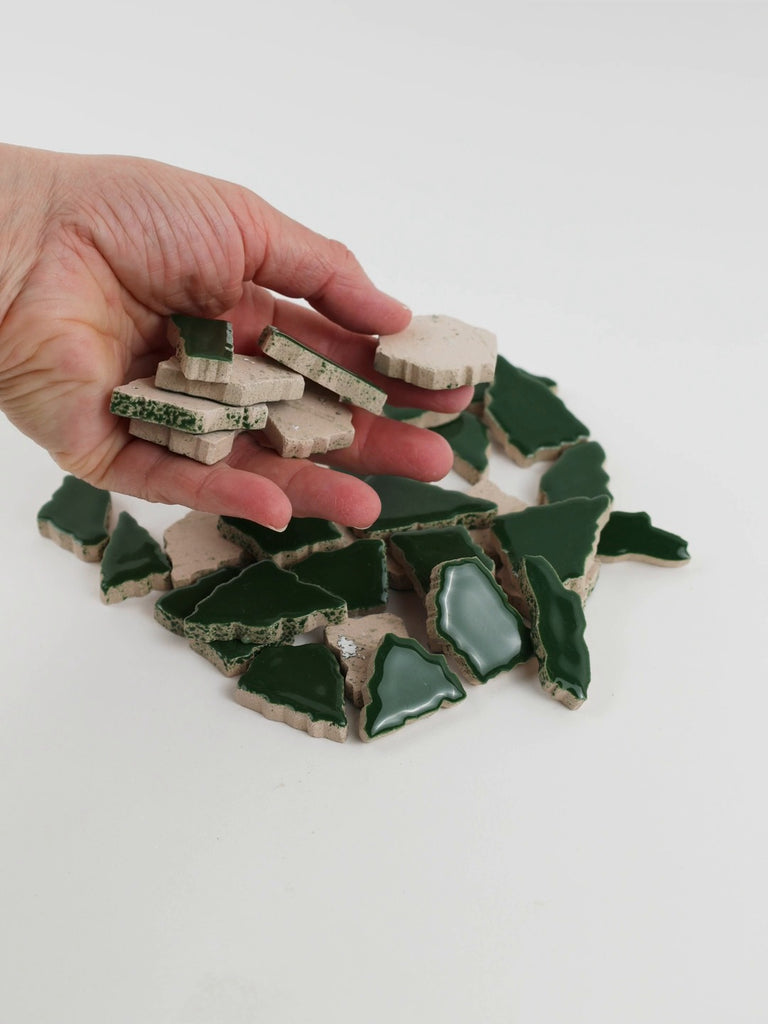 Video of Flip XL ceramic puzzle pieces in moss green, opaque glazed porcelain, 20–60 mm, picked up from a pile by hand and dropped back to show their size, shapes and finish