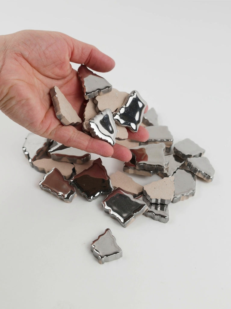 Video of Flip XL ceramic puzzle pieces in silver, opaque glazed porcelain, 20–60 mm, picked up from a pile by hand and dropped back to show their size, shapes and finish